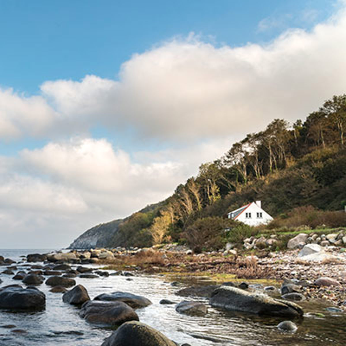 Hvidt hus beliggende ved skovklædt kystlinje med havudsigt på Bornholm under en overskyet himmel.