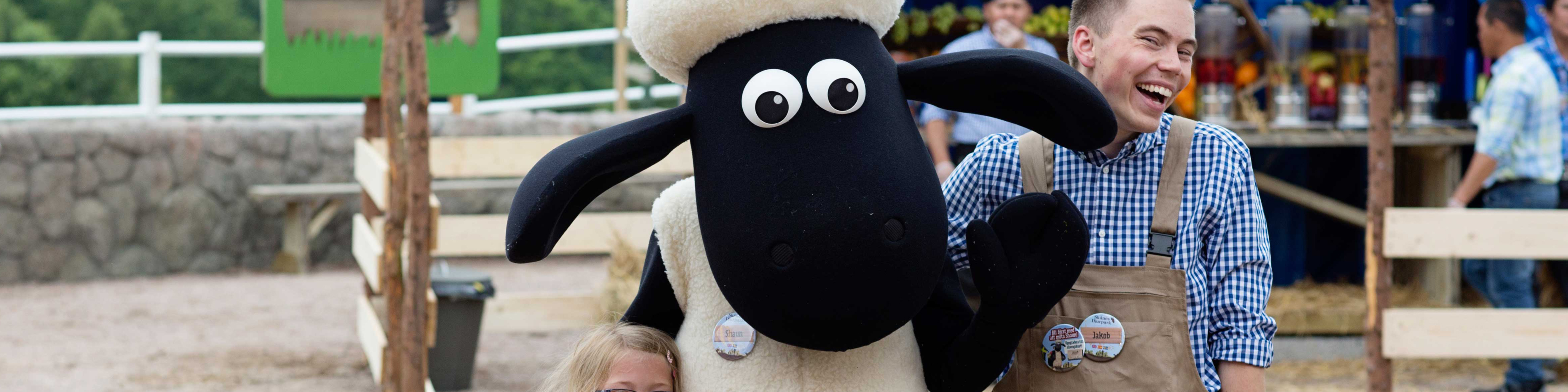 Shaun fåret krammer en glad mand i ternet skjorte, omgivet af en udendørs markedsplads.