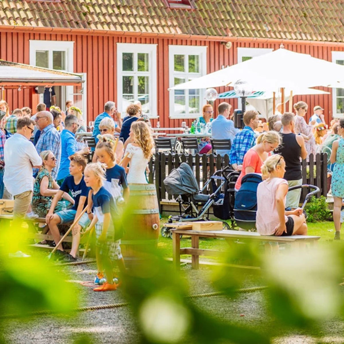 Folk samles til en udendørs sommerfest foran et rødt træhus under en solrig himmel.