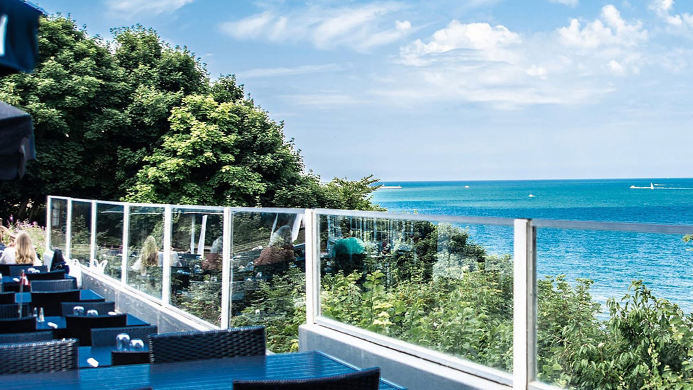 Modern terrass med glasräcke Fyrkroen och havsutsikt, omgiven av frodigt grönt landskap och klarblå himmel.