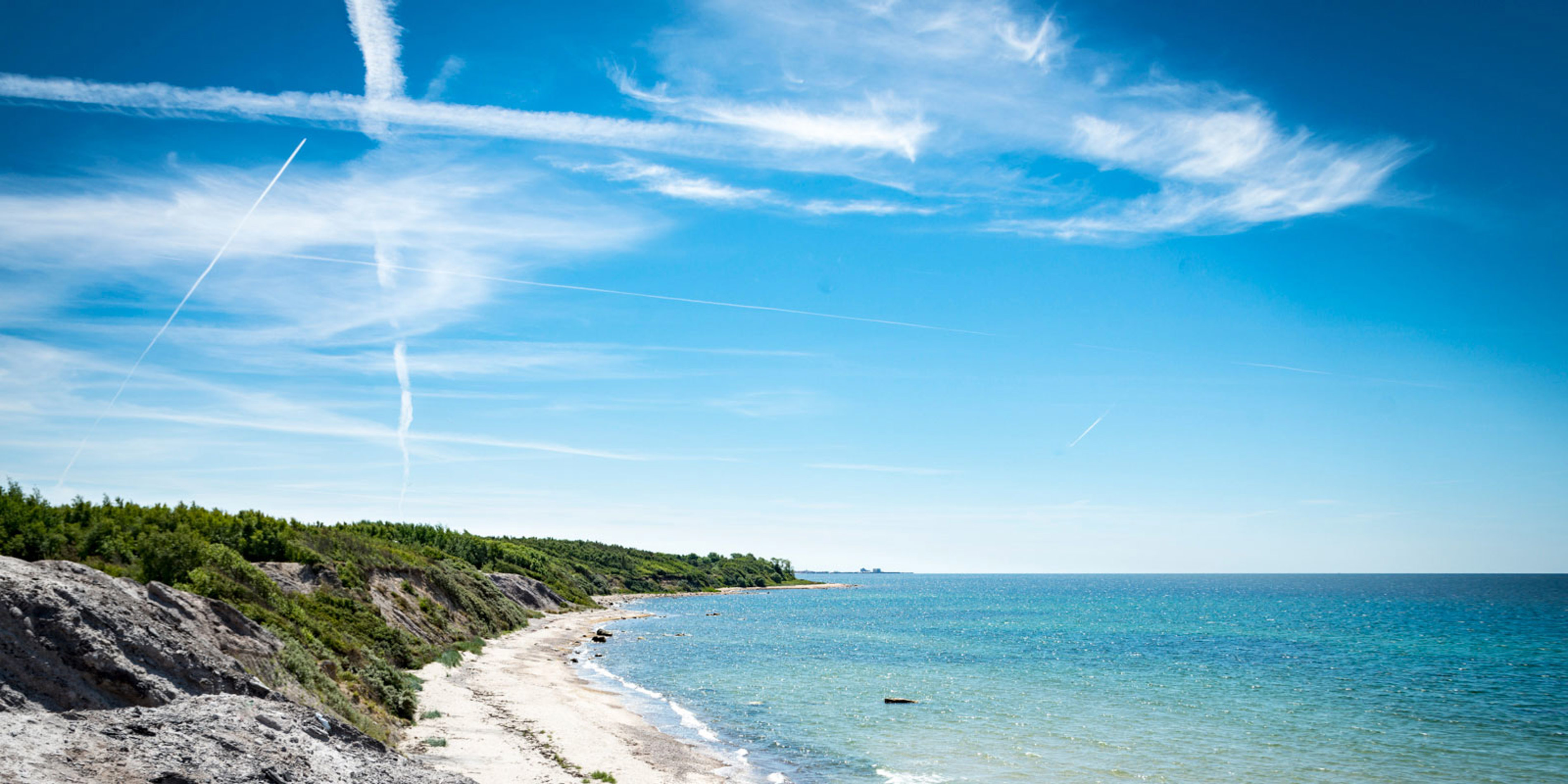 Küstenlinie mit Strand, Wald und klarem, blauem Wasser unter sonnigem, blauem Himmel.
