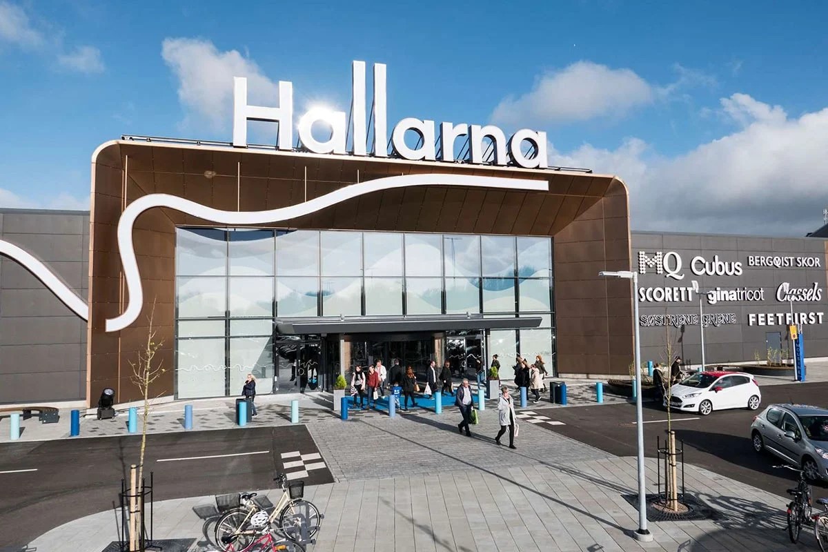 Indgang til shoppingcenteret Hallarna med kunder der går ind og ud under det store skilt på solrig dag.