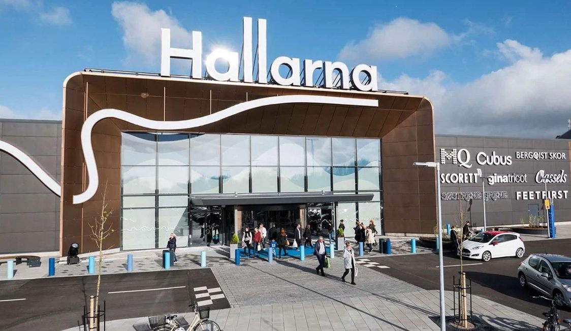 Indgang til shoppingcenteret Hallarna med kunder der går ind og ud under det store skilt på solrig dag.