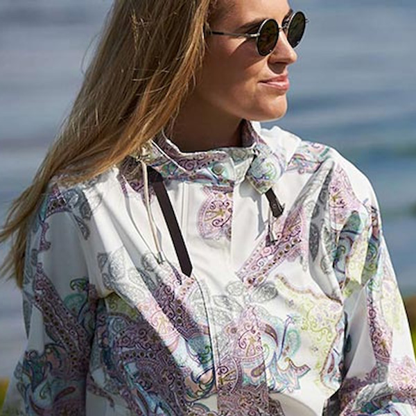 Ilse Jacobsen regnkappa för kvinnor i vitt med färgglad paisleymönster, bärs av kvinna vid kust.