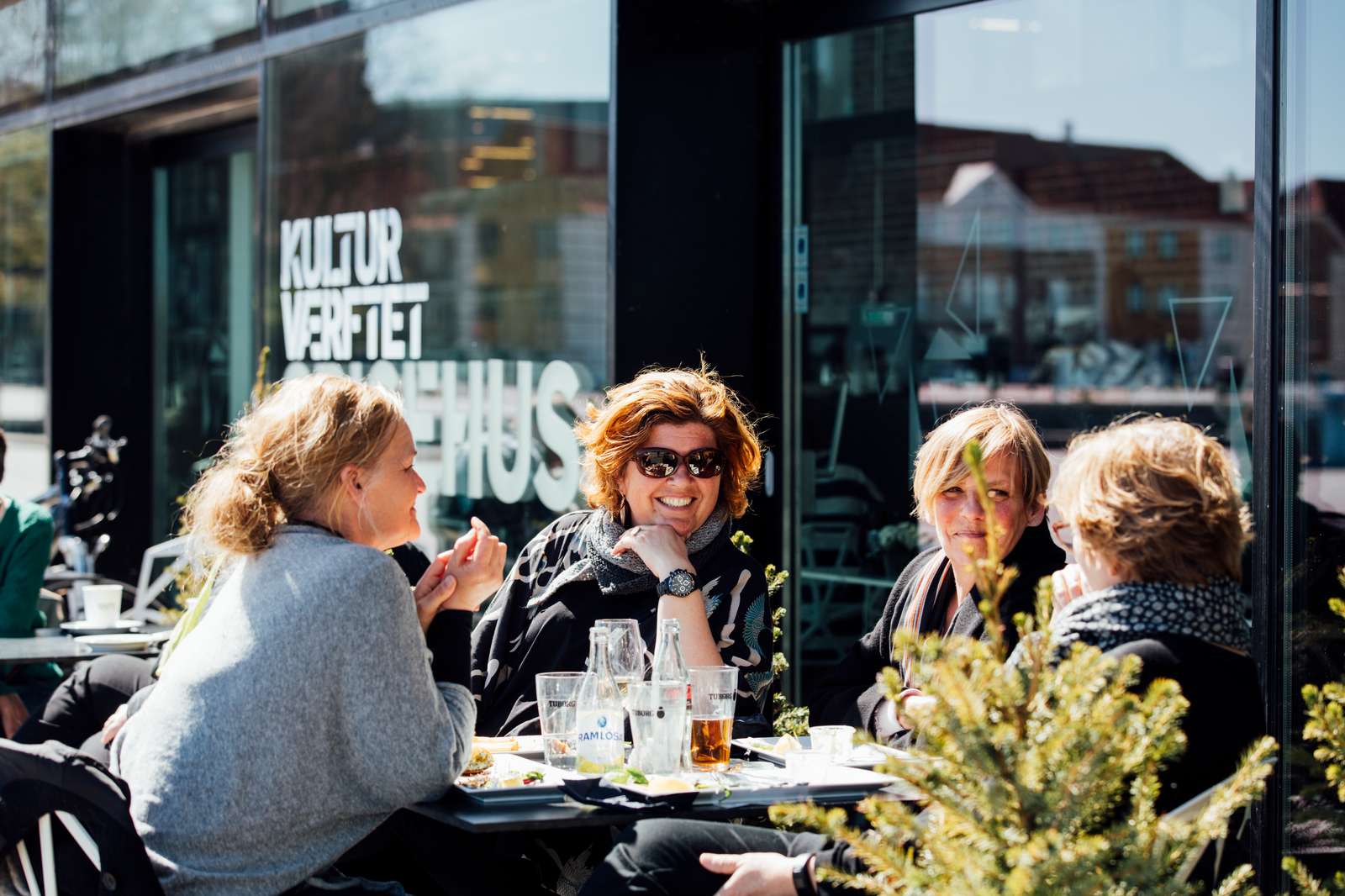 Människor njuter av en solig dag på ett kafé, med glas i förgrunden och reflekterande fönster i bakgrunden.