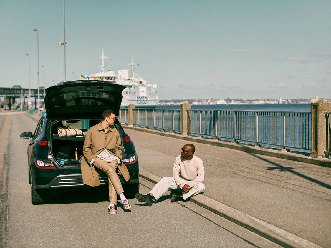 Två män vid en bil med öppen baklucka, som pratar och njuter av en solig dag vid havet.