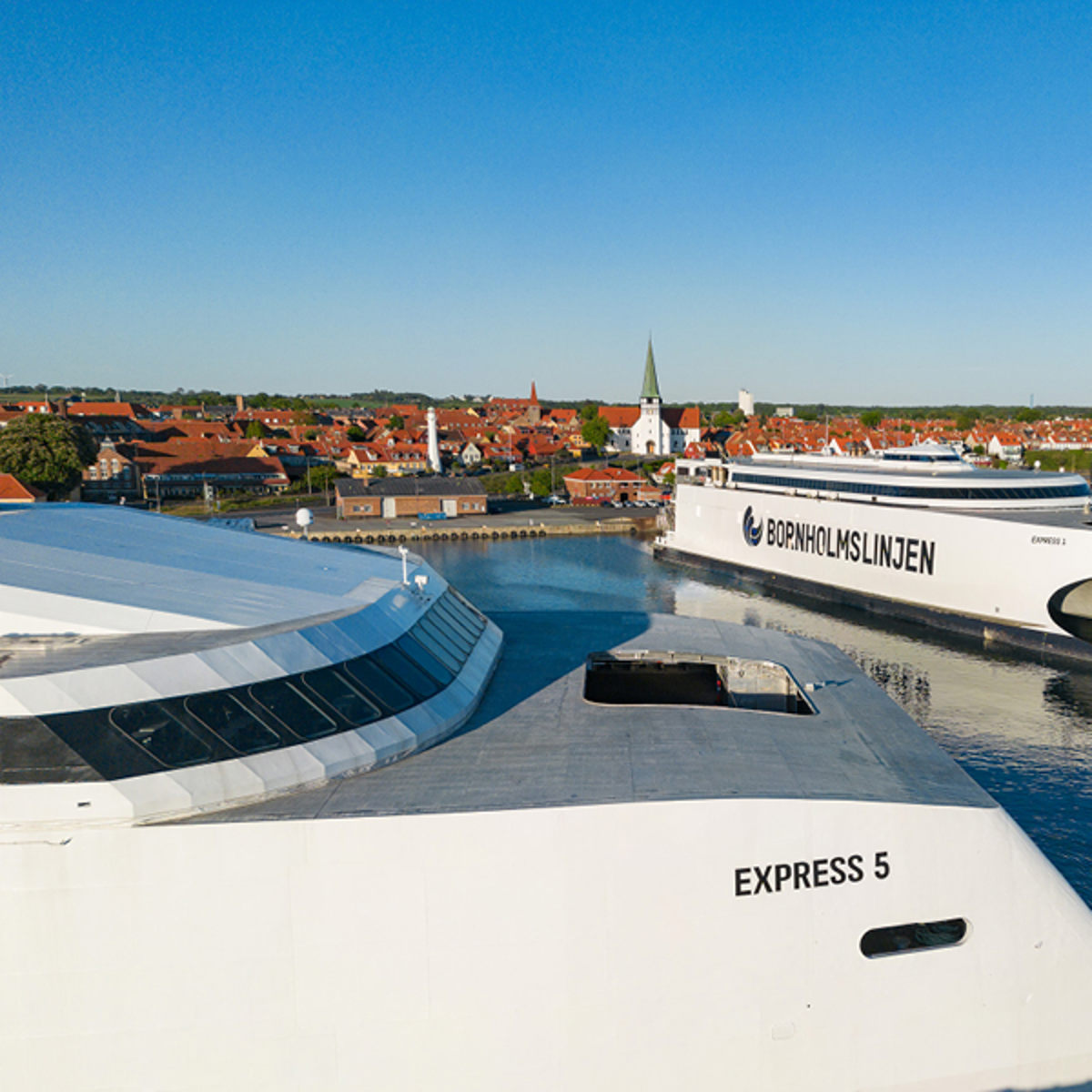 Bornholmslinjen færge Express 5 nærmer sig havn i solskin, med spæren af byens røde tage i baggrunden.