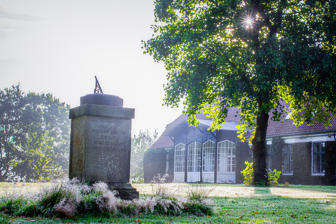 Solur på en gammel stenbase i en solrig have ved Rungstedlund med træ og bygning i baggrunden.
