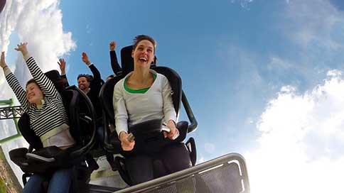 Kvinde i rutsjebane griner med hænderne oppe mod en blå himmel under en spændende tur i forlystelsespark.
