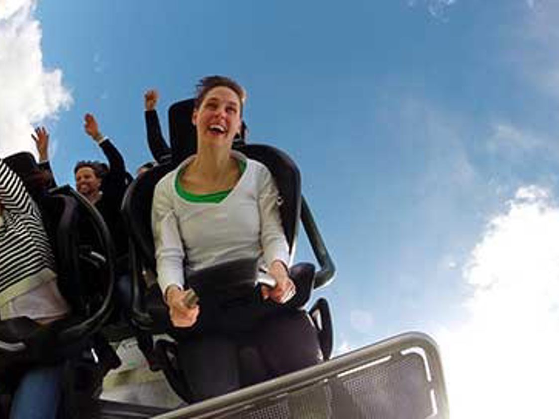 Kvinde i rutsjebane griner med hænderne oppe mod en blå himmel under en spændende tur i forlystelsespark.