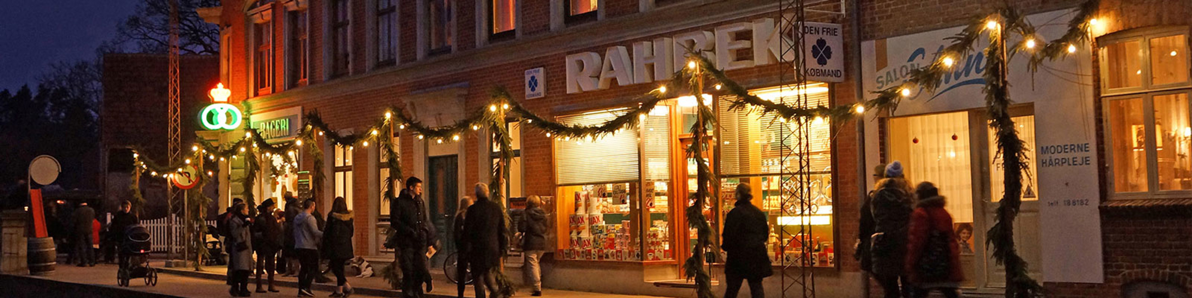 Kosmopolitisk scene om aftenen ved julepyntet forretning med skiltet 'RAHBEK', mennesker går forbi.