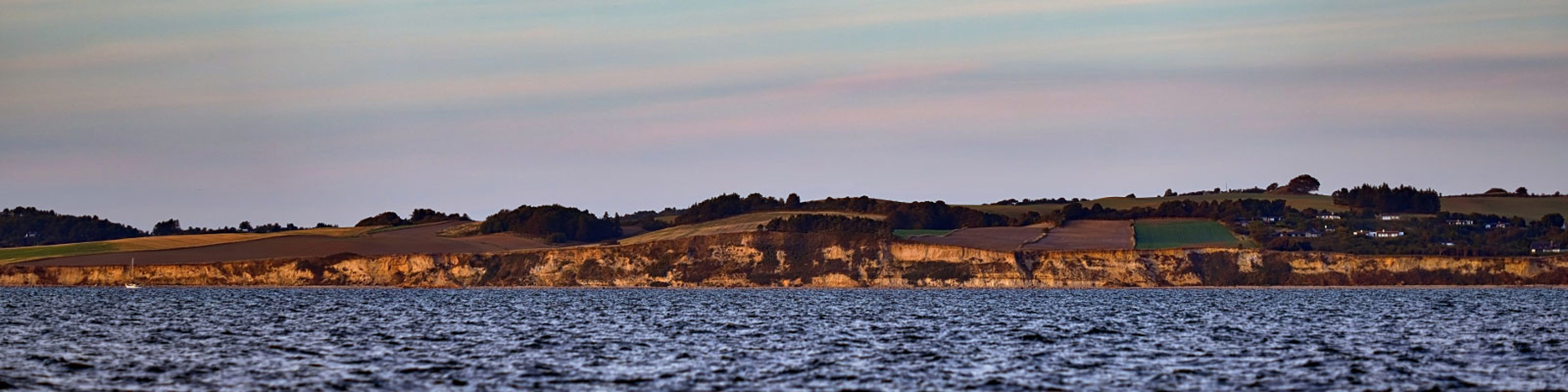 Panoramisk udsigt over Mols Bjerge med skrænter og landskab fra havet i solnedgangsbelysning.