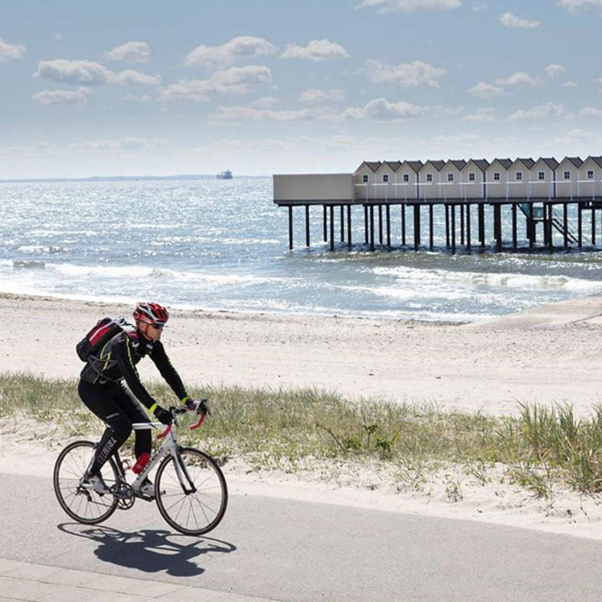 Cyklist i cykeltøj og hjelm kører på landevej ved stranden med havet og huse på pæle i baggrunden.