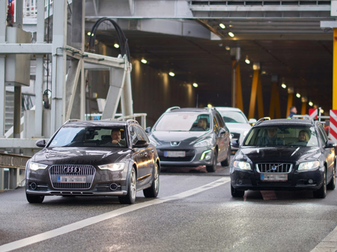 Biler kører af færgen efter ankomst til havn, med fokus på en sort bil på vej ud af færgen i tæt trafik.