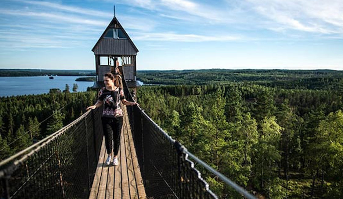 Kvinde går på hængebro mod udkigstårn i skovområde med udsigt over vandet og skovlandskap.