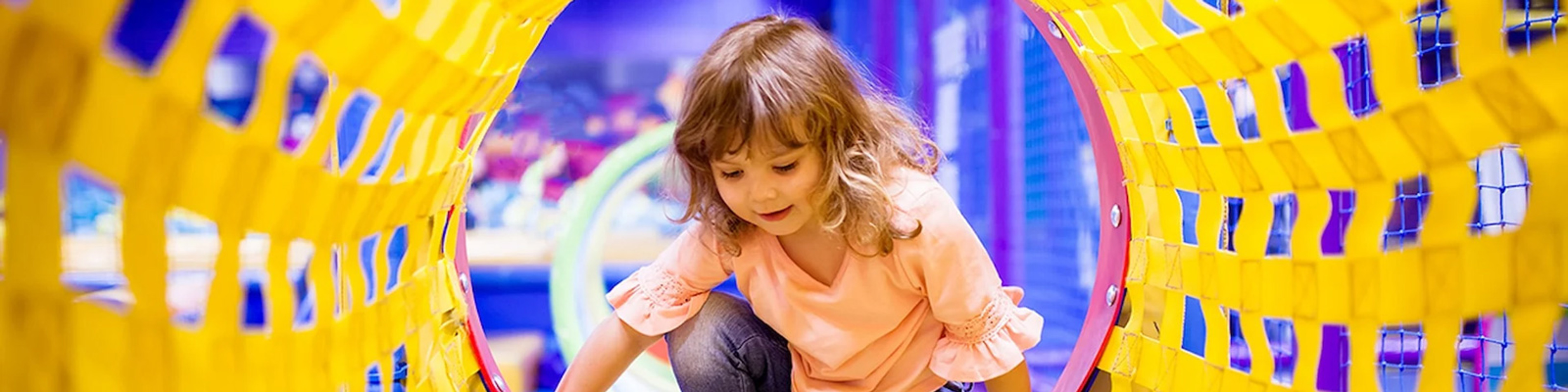 Lille pige i lyserød T-shirt leger i et farverigt, gult klatrerør i en indendørs legeplads.