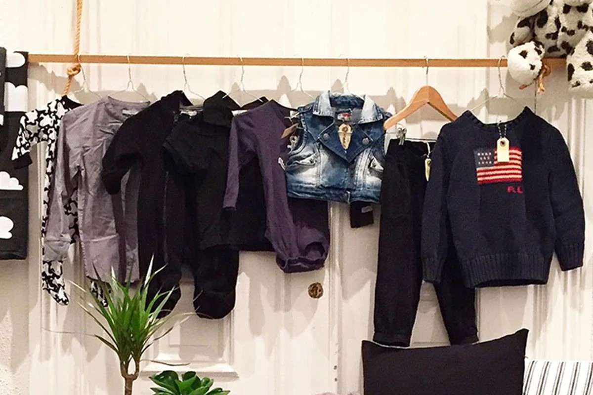 Børnetøj i forskellige farver hænger på en træbøjle mod hvid baggrund. Denimvest, lilla og sort bluser ses tydeligt.