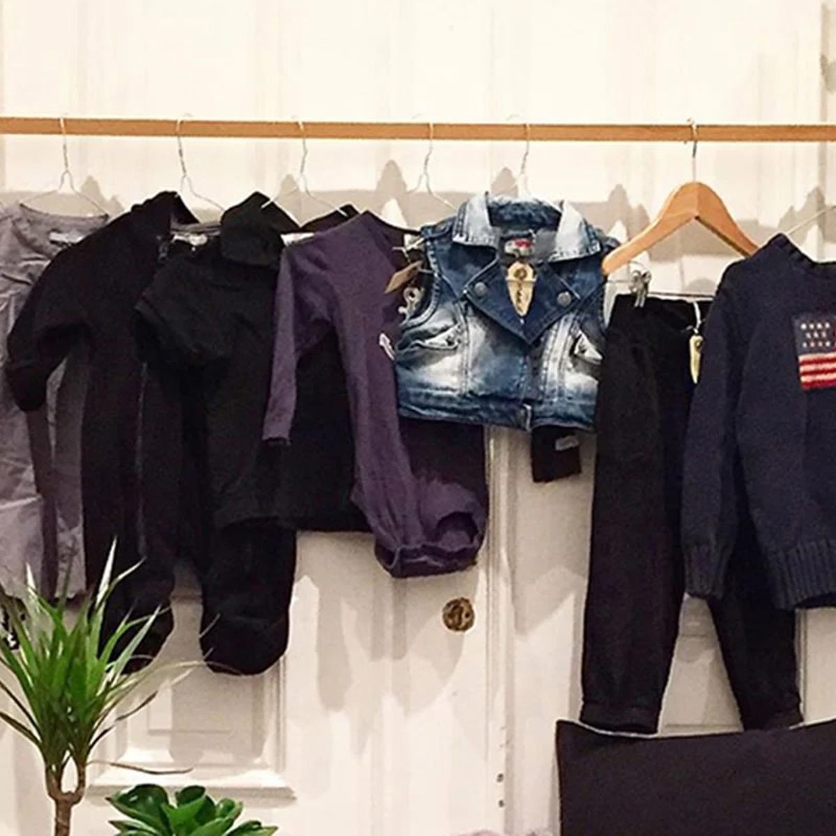 Børnetøj i forskellige farver hænger på en træbøjle mod hvid baggrund. Denimvest, lilla og sort bluser ses tydeligt.