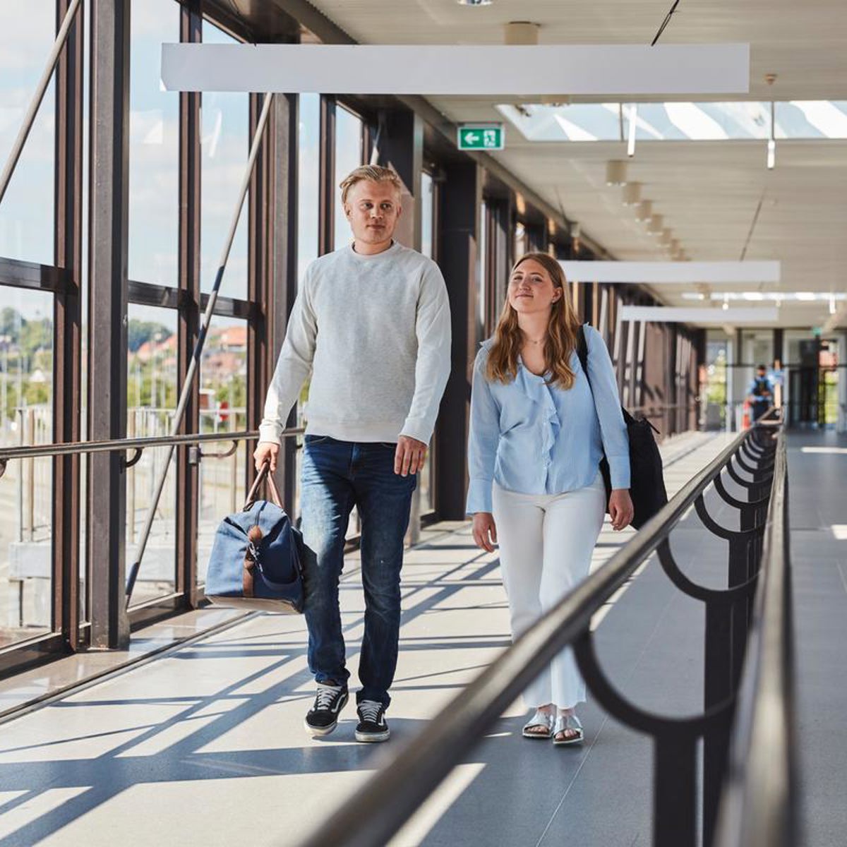 Ungt par iført afslappet tøj går gennem en moderne gangbro i lufthavnsbygning.