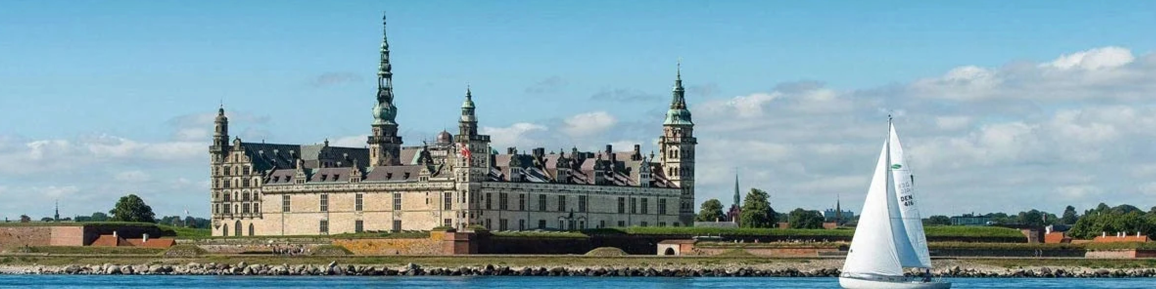 Kronborg Slot set fra havet med en sejlbåd i forgrunden under en klar blå himmel.