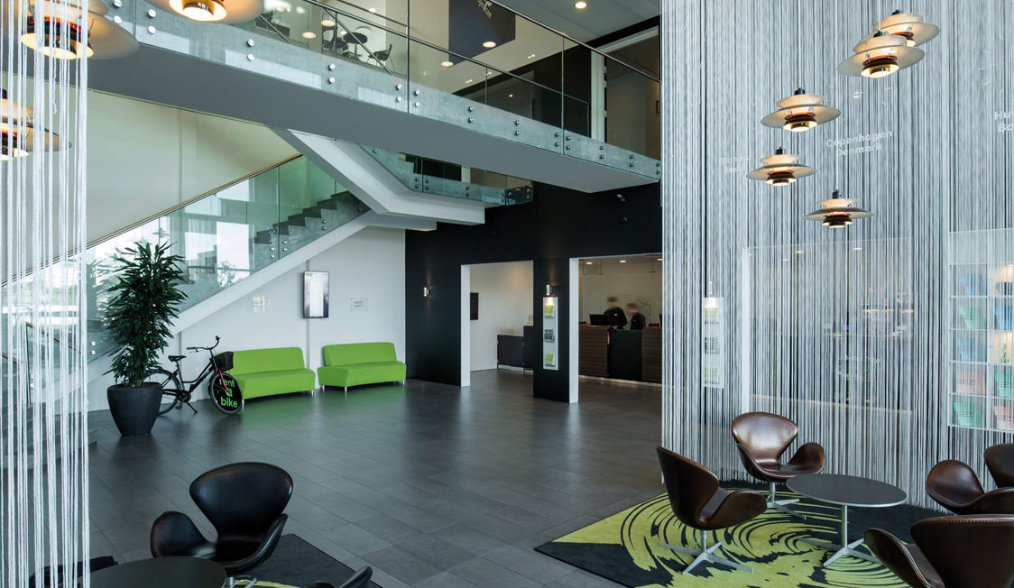 Moderne kontorlobby med grønne sofaer og glasgelænder set fra siden ved receptionen.