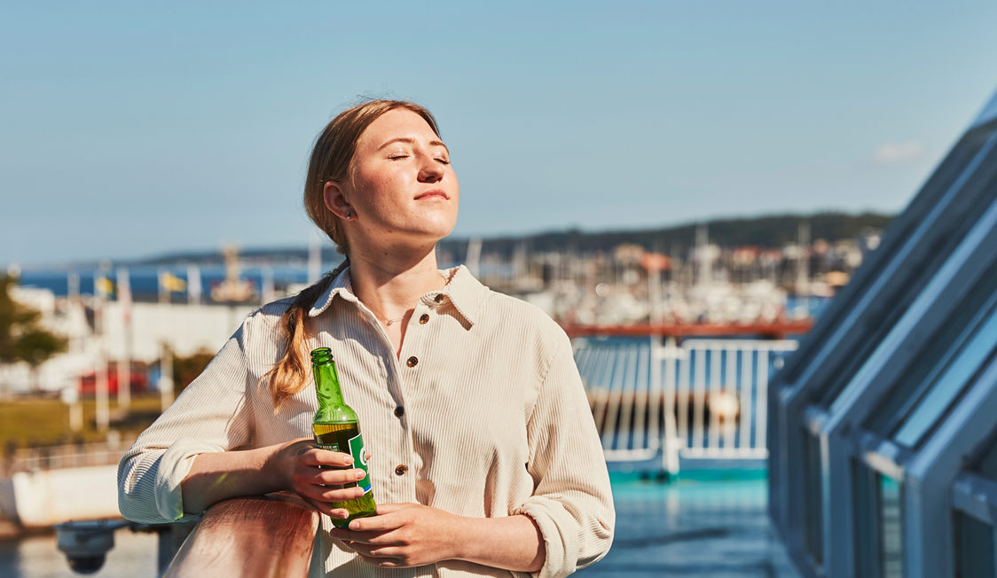 Kvinde i beige skjorte nyder solen på færgen med en flaske i hånden, omgivet af en klar blå himmel.