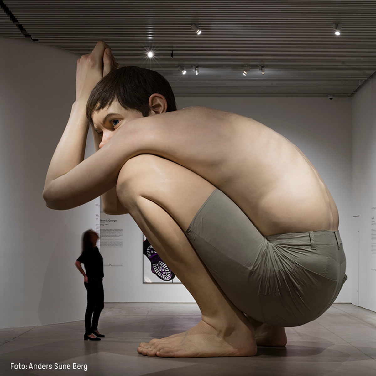 Gigantisk skulptur af knælende dreng i brunlige shorts udstillet i kunstgalleri.