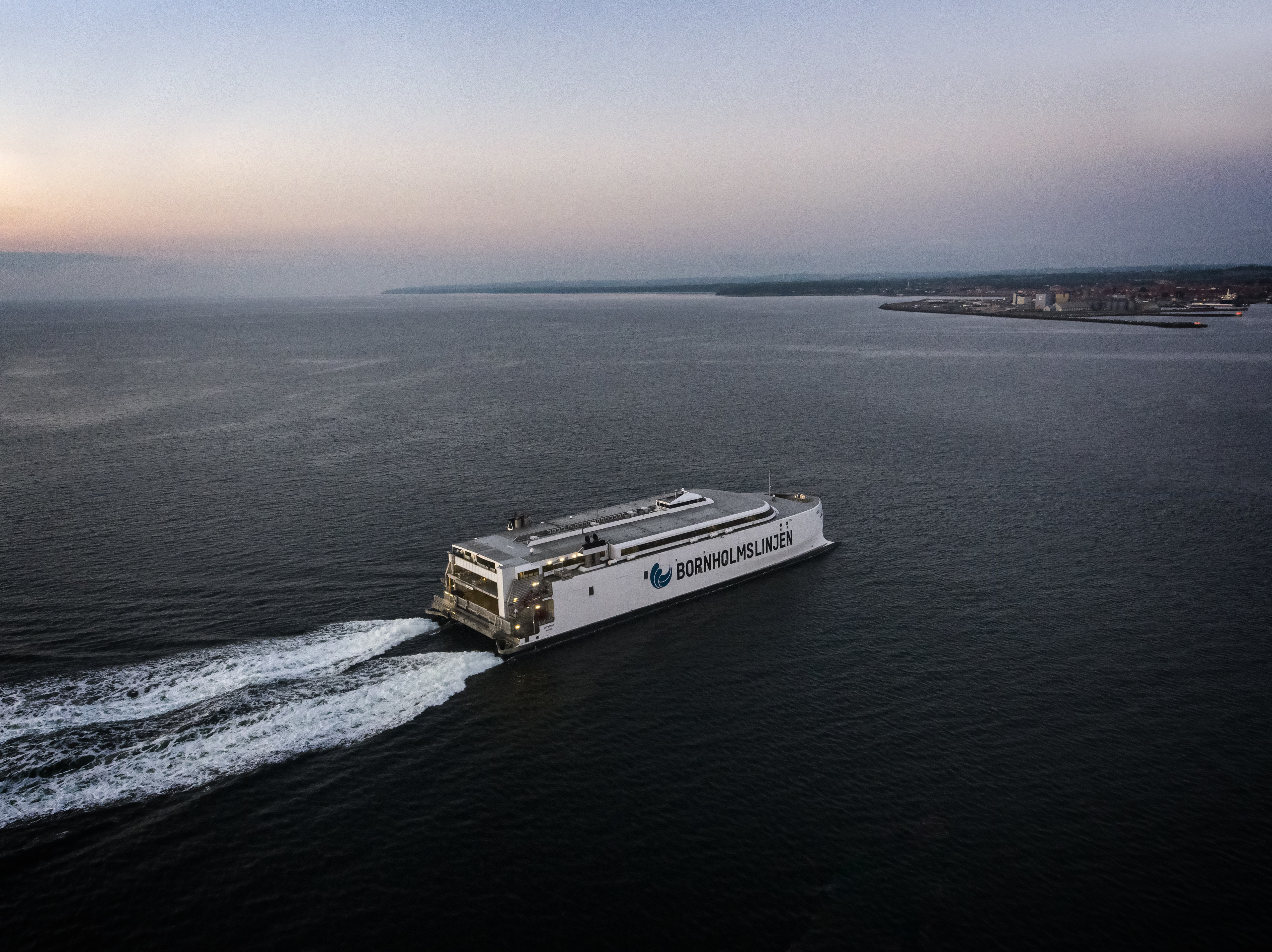 Bjørnlinjen færge sejler på åbent hav mod horisonten ved solopgang i fugleperspektiv.