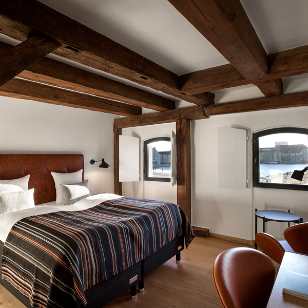 Komfortabelt hotelværelse med træbjælker, stor seng med stribet sengetæppe og udsigt over vandet gennem vinduerne.