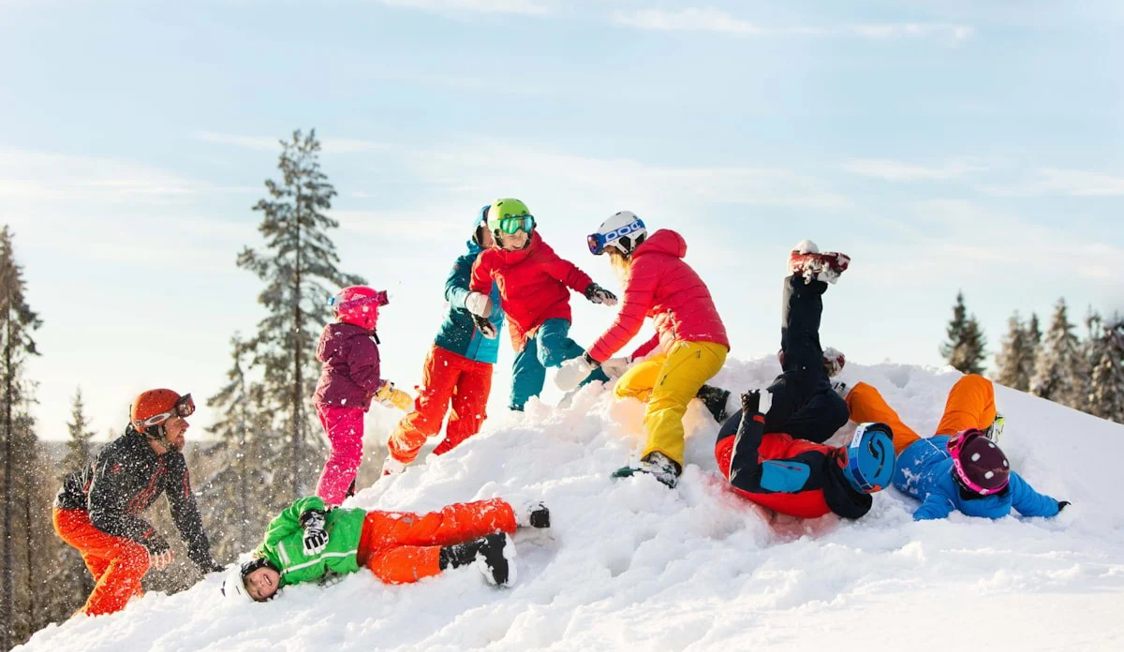 Børn i farverigt skitøj leger og ruller i sneen på en bakke på en klar vinterdag.