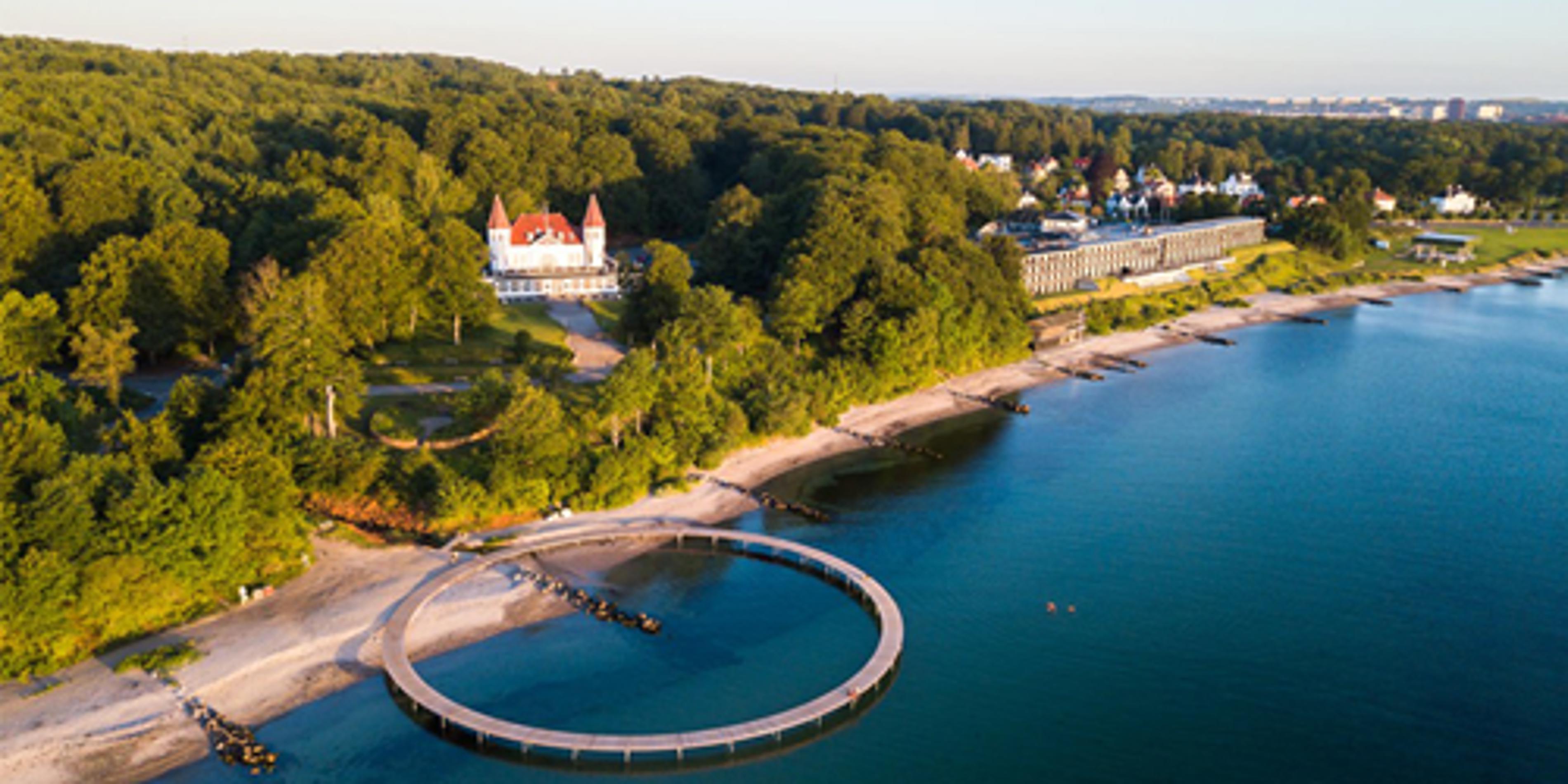 Cirkulær badebro ved skovklædt kystlinje i Vejle med udsigt til moderne villa og blå hav i fugleperspektiv.