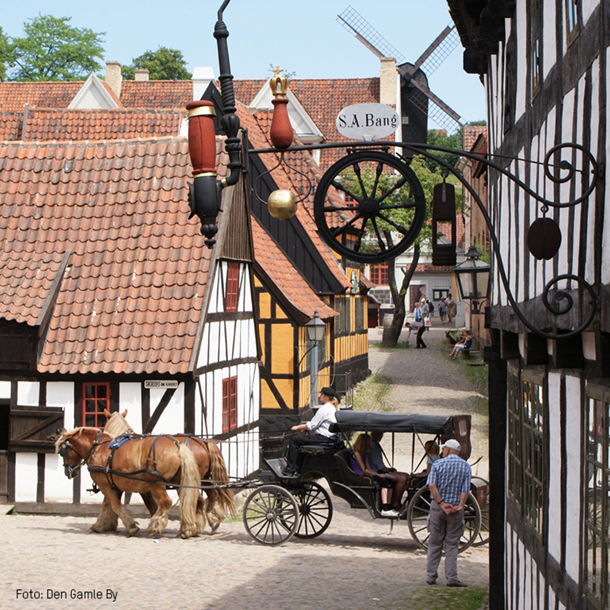 Hestevogn kører gennem historisk landsby i Den Gamle By med bindingsværkshuse og mølle i baggrunden.