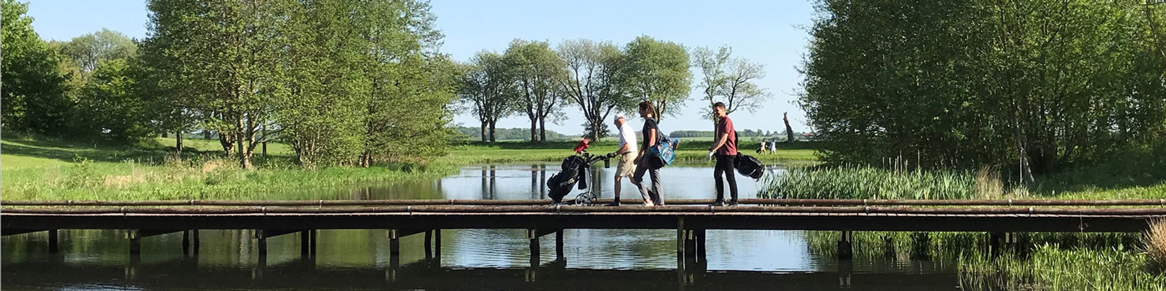 Folk, der går over en træbro med golfudstyr, omkranset af grønt landskab og blå himmel.