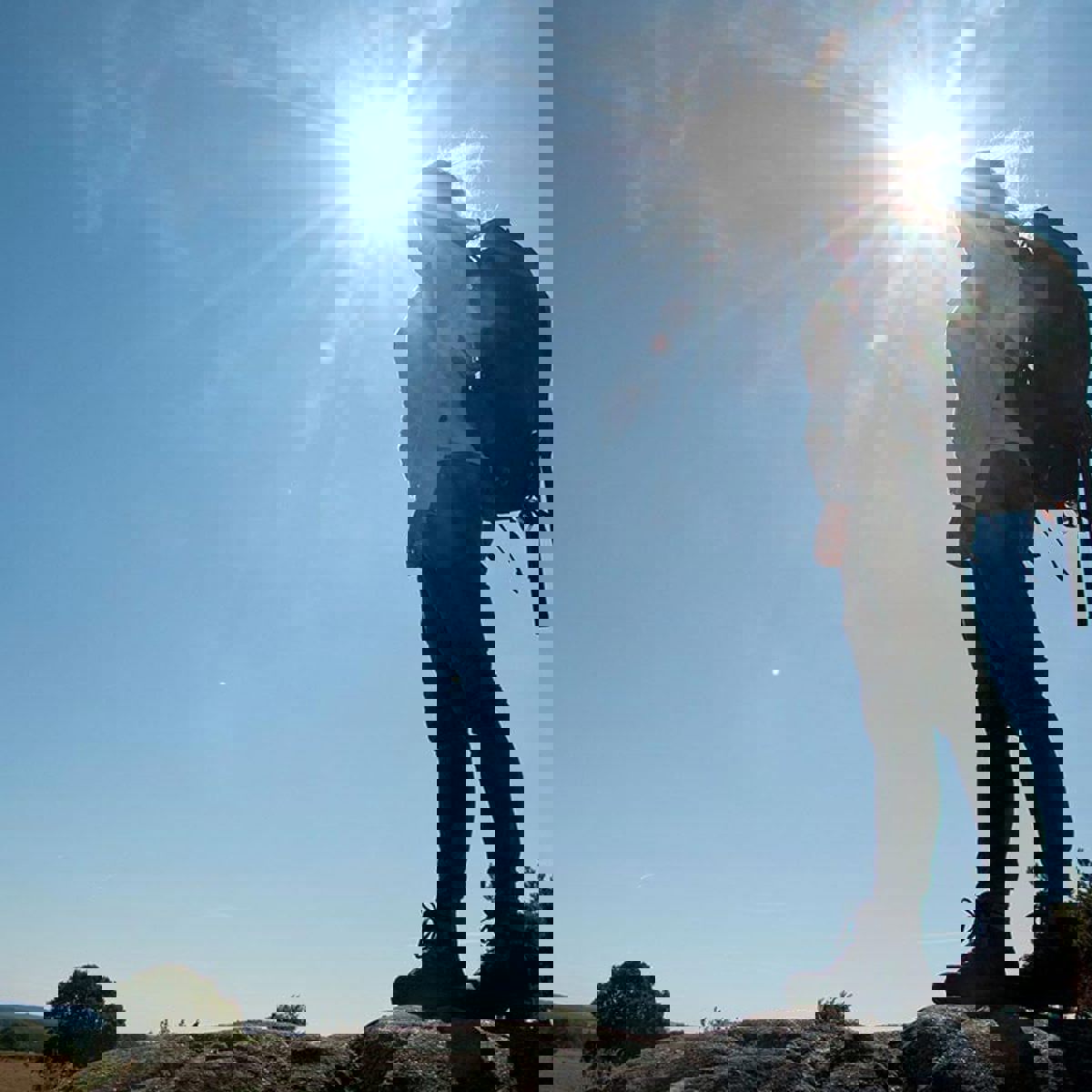 En person ses fra frøperspektiv stå i solskin på en vandretur med rygsæk.