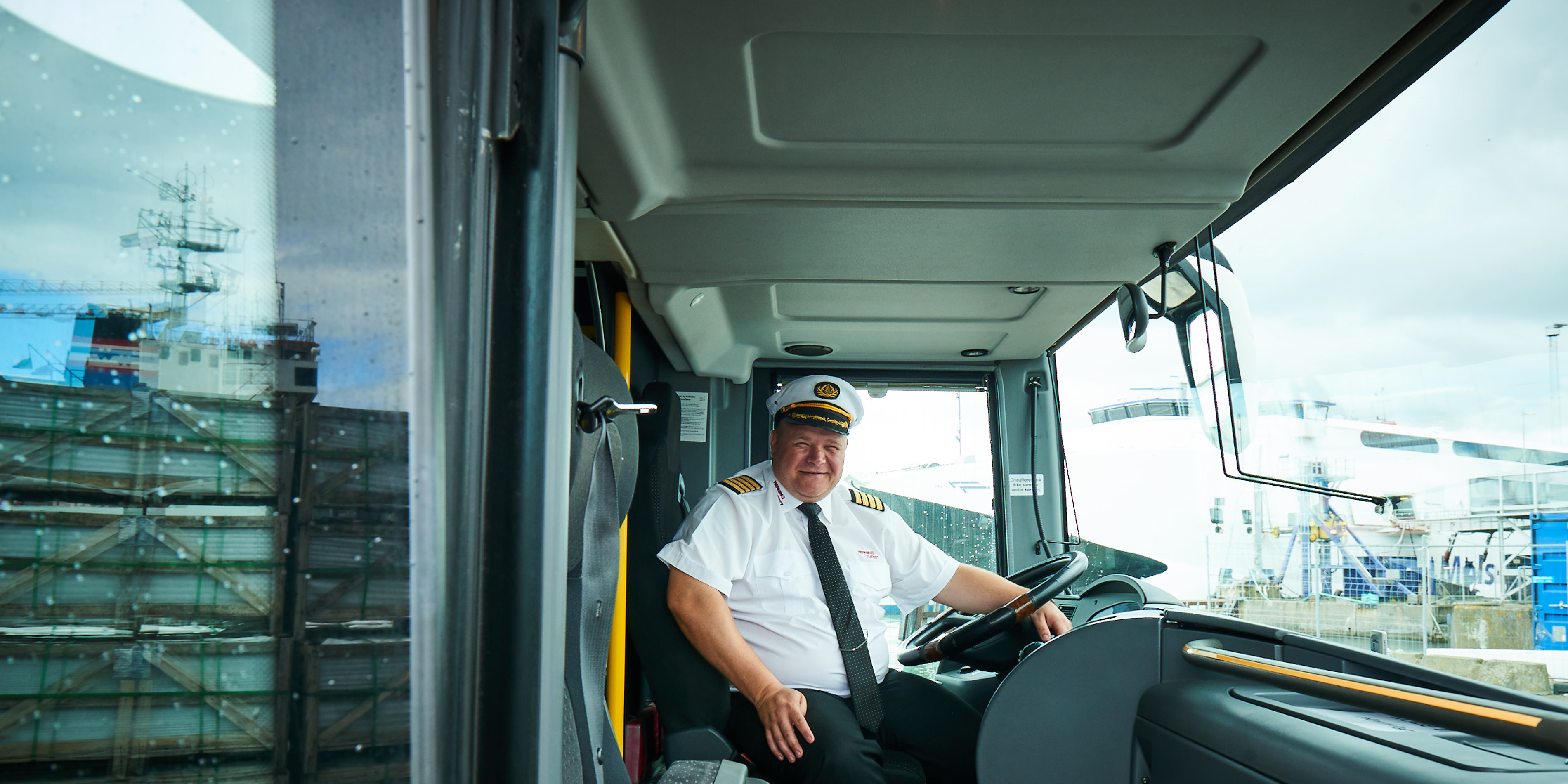 Buschauffør iført uniform sidder bag rattet i en moderne bus med udsigt til en færge gennem forruden.