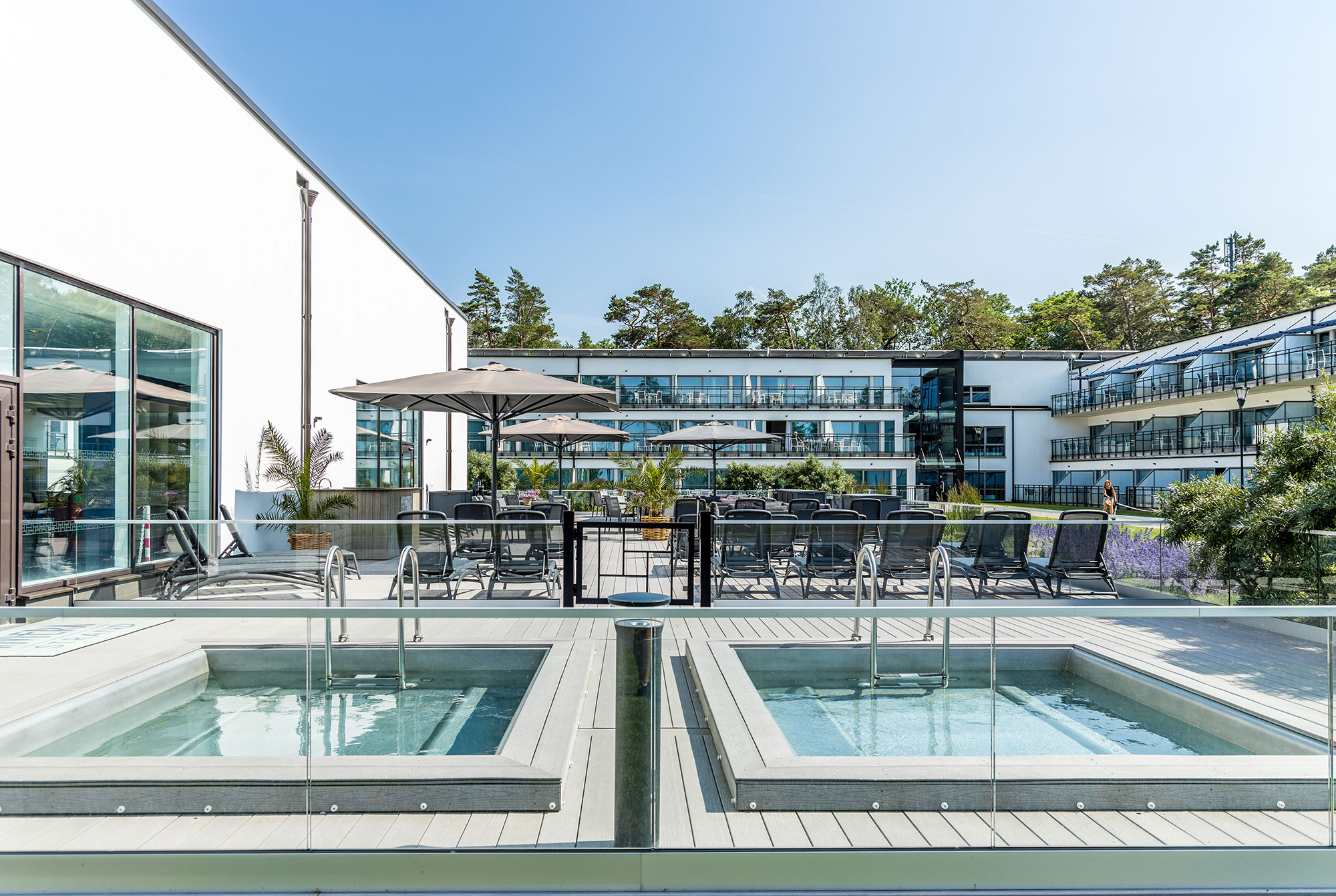 Udendørs terrasse med to spabade, liggestole og parasoller foran et moderne hotel i naturskønne omgivelser.