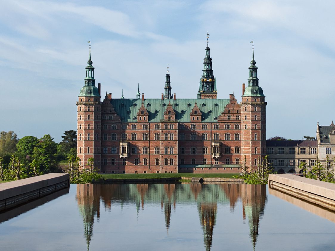 Frederiksborg Slot reflekteres i søen på en solrig dag med klar blå himmel.