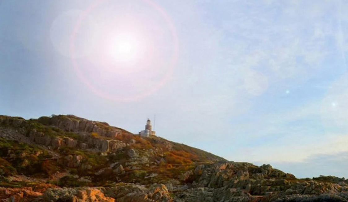Fyrtårn på en klippe med den klare himmel og solens stråler over landskabet i horisonten.