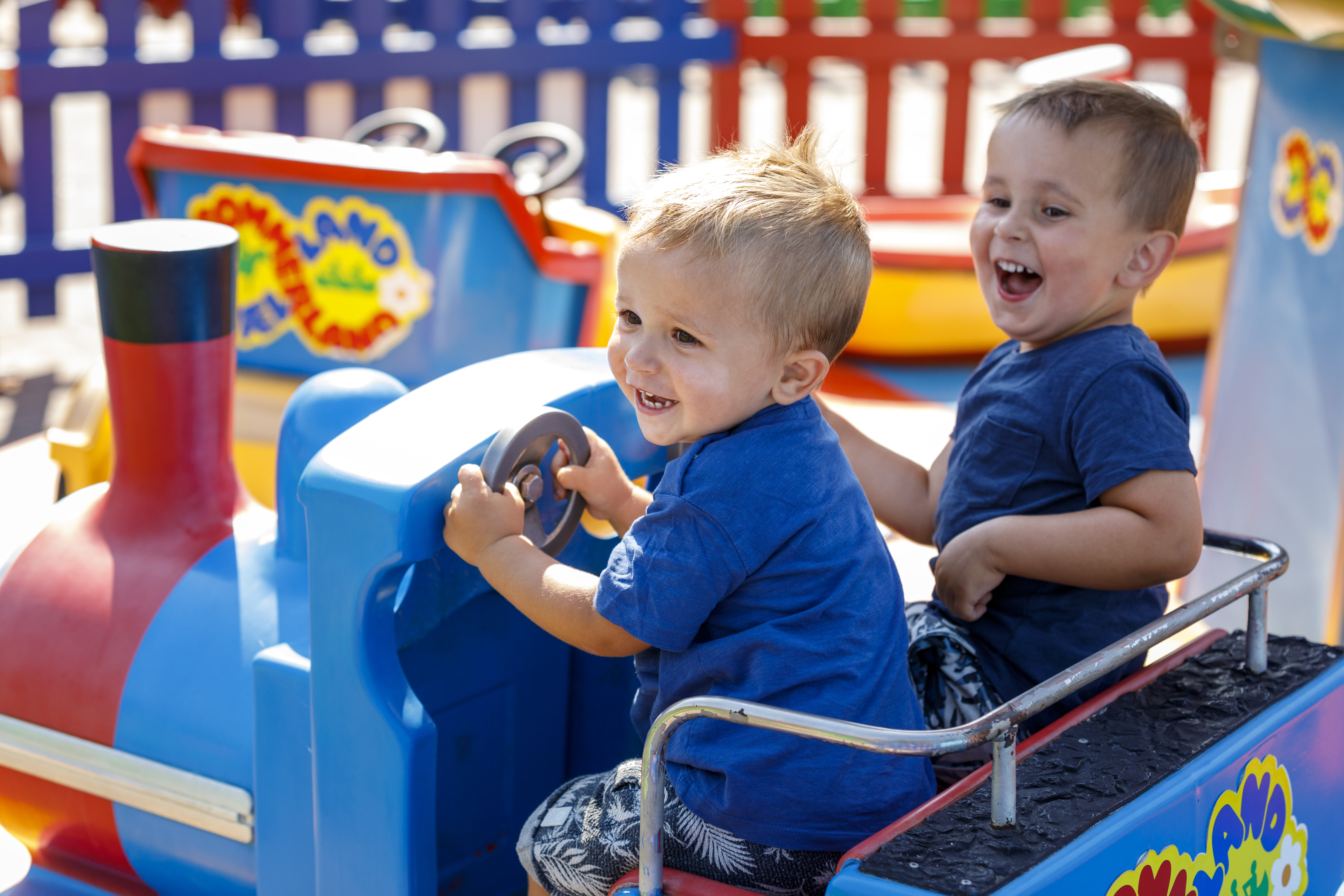 Små barn skrattar och kör en blå leksaksbil på en färgglad lekplats under en solig dag.