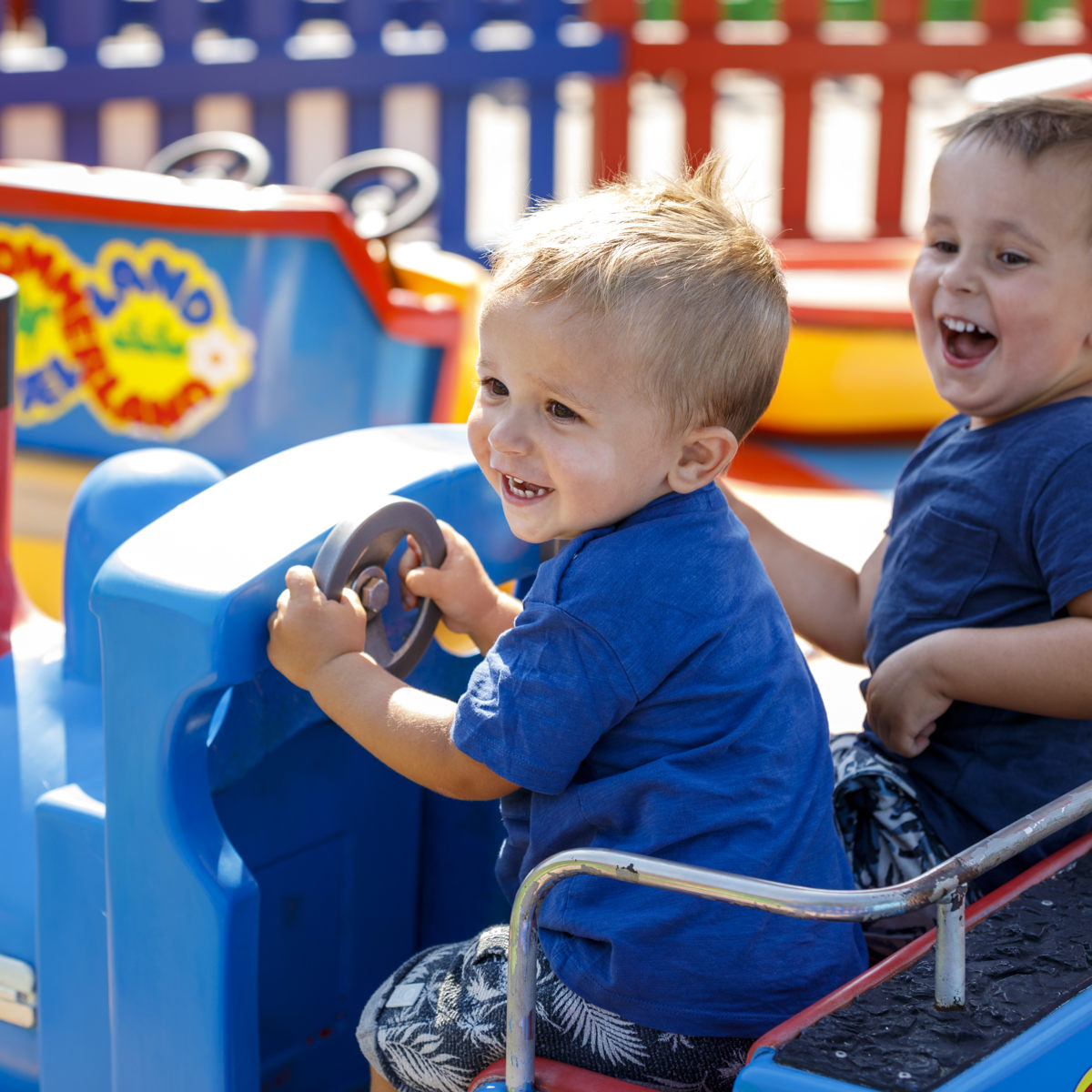 Små barn skrattar och kör en blå leksaksbil på en färgglad lekplats under en solig dag.