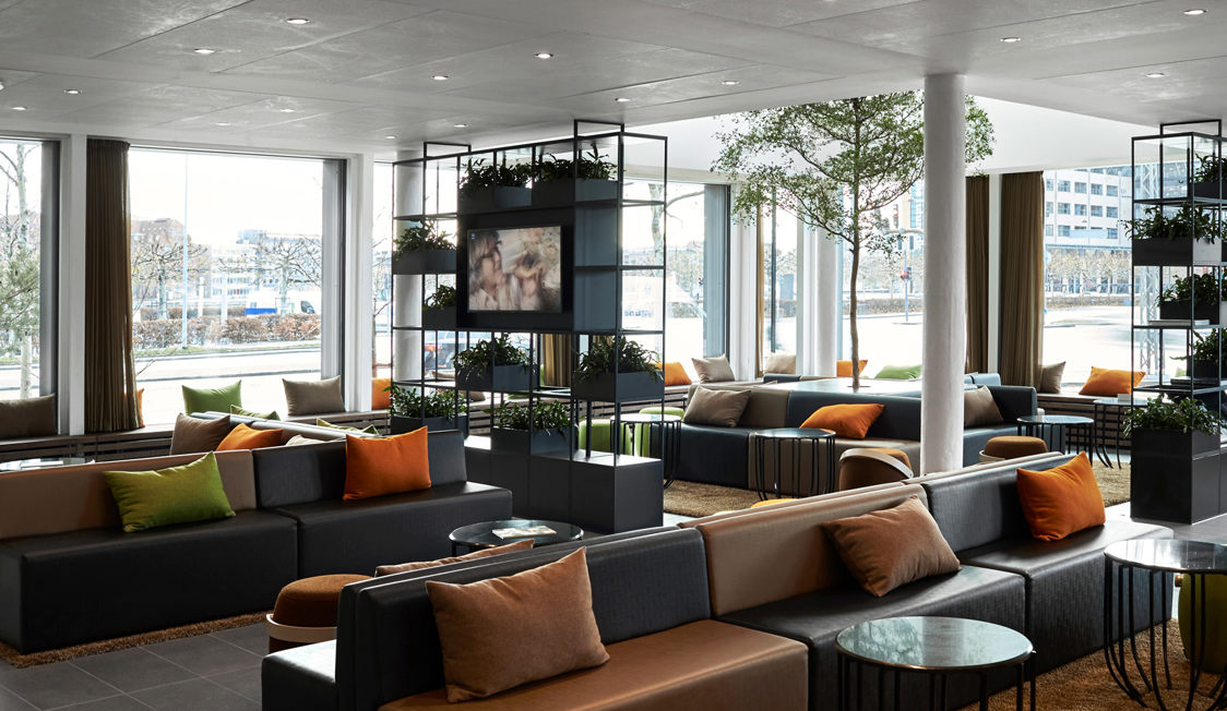 Moderne lounge med grå sofaer og farverige puder, omgivet af store vinduer og grønne planter.