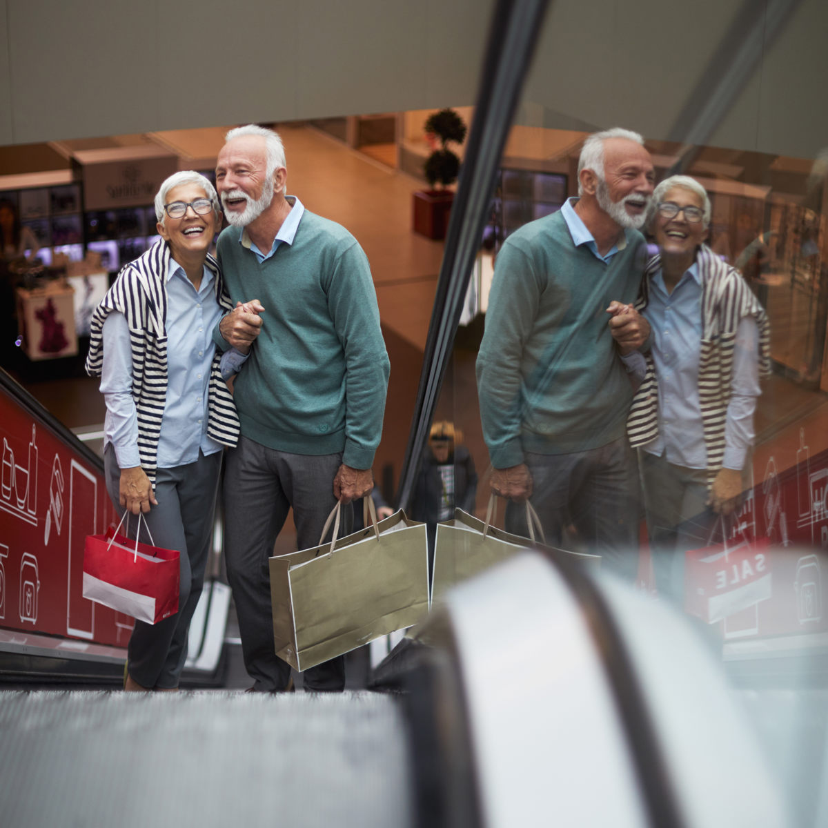 Ældre par på rulletrappe i indkøbscenter, griner og holder indkøbsposer og hinandens hænder.