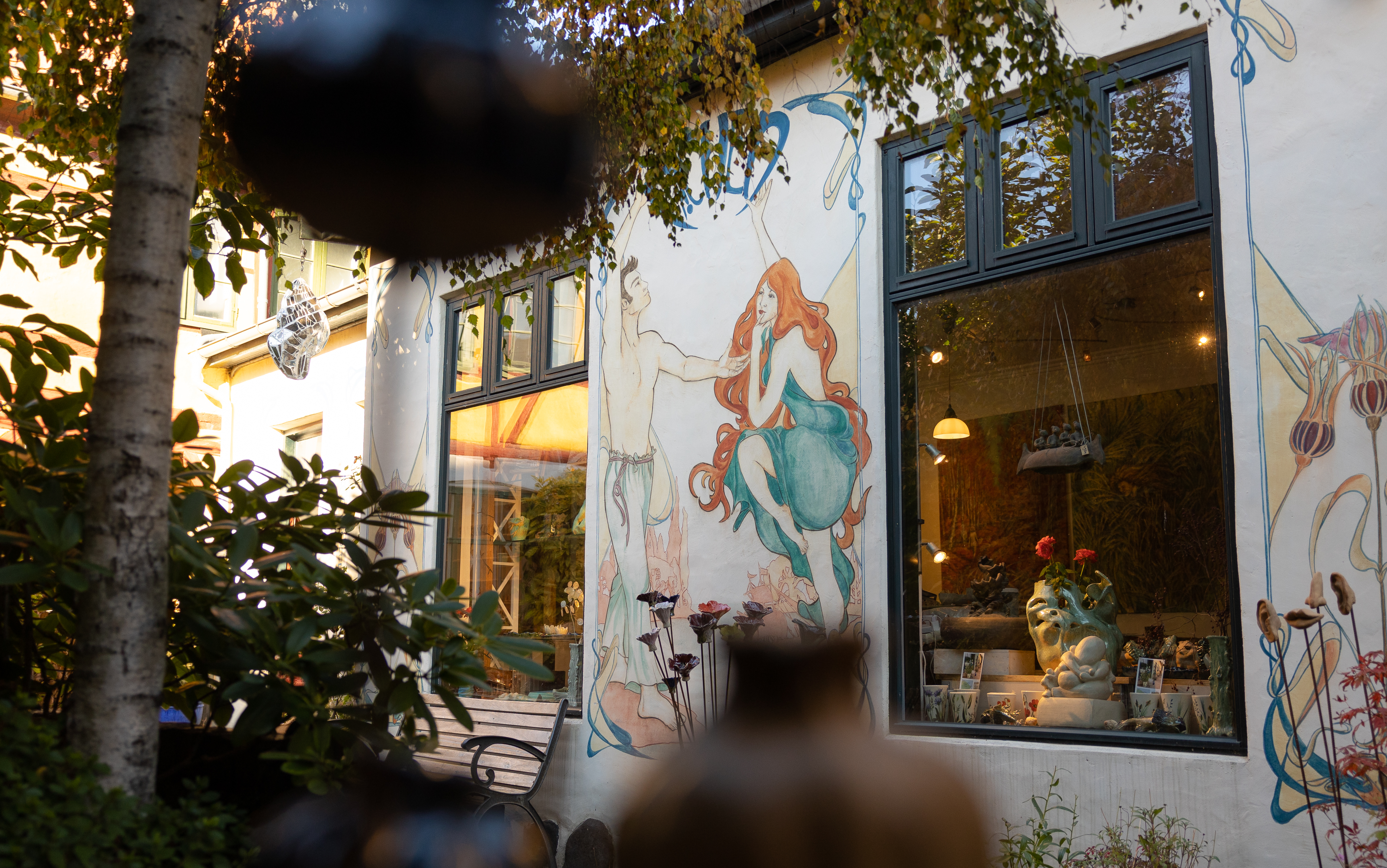 Väggmålning med en kvinna och man i livlig dialog, omgiven av grönska och synligt fönster med keramik i bakgrunden.