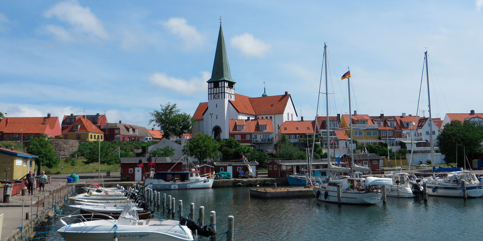 Kirche in Bornholm mit markantem grünen Turm, umgeben von Segelbooten und bunten alten Häusern entlang des Hafens.