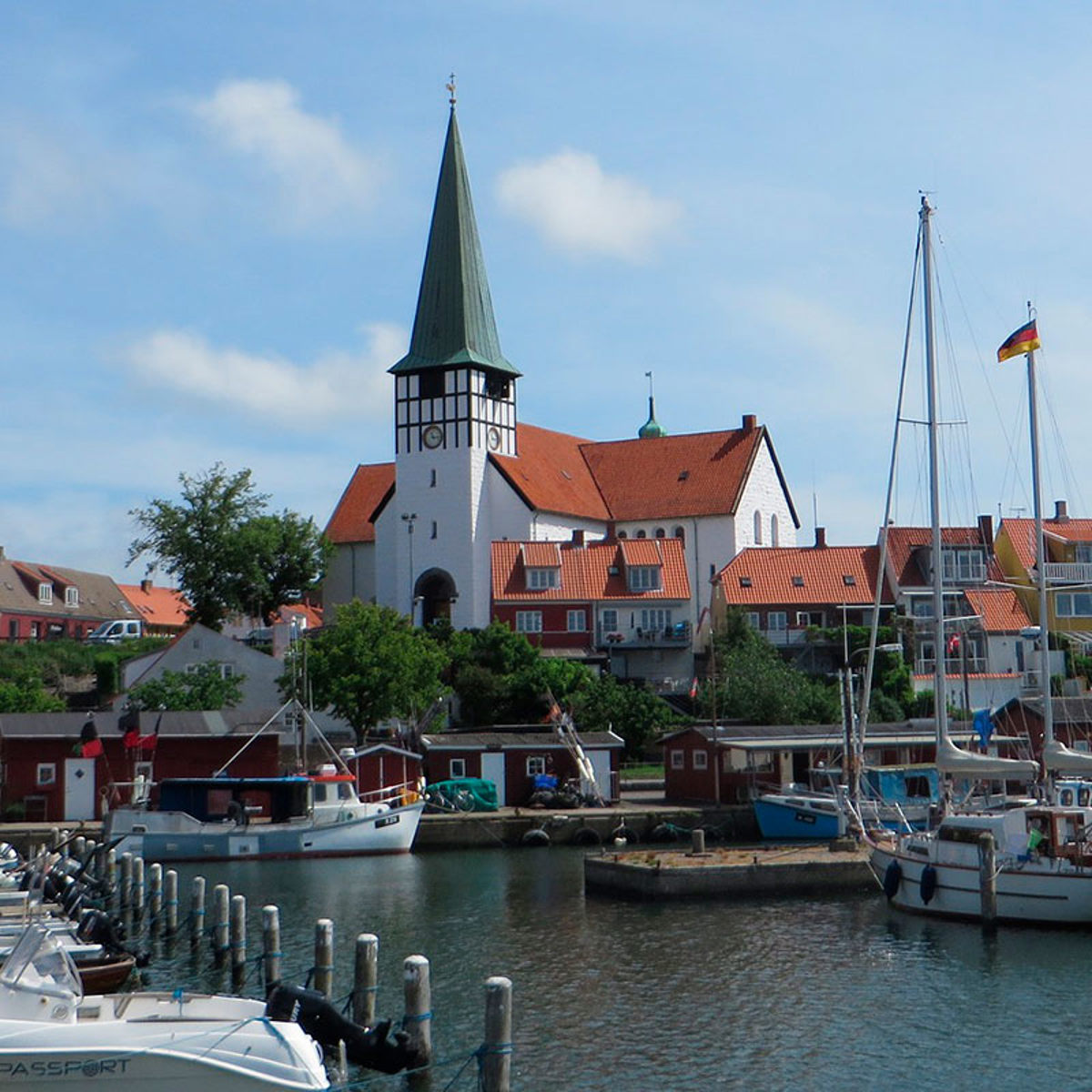 Kirche in Bornholm mit markantem grünen Turm, umgeben von Segelbooten und bunten alten Häusern entlang des Hafens.