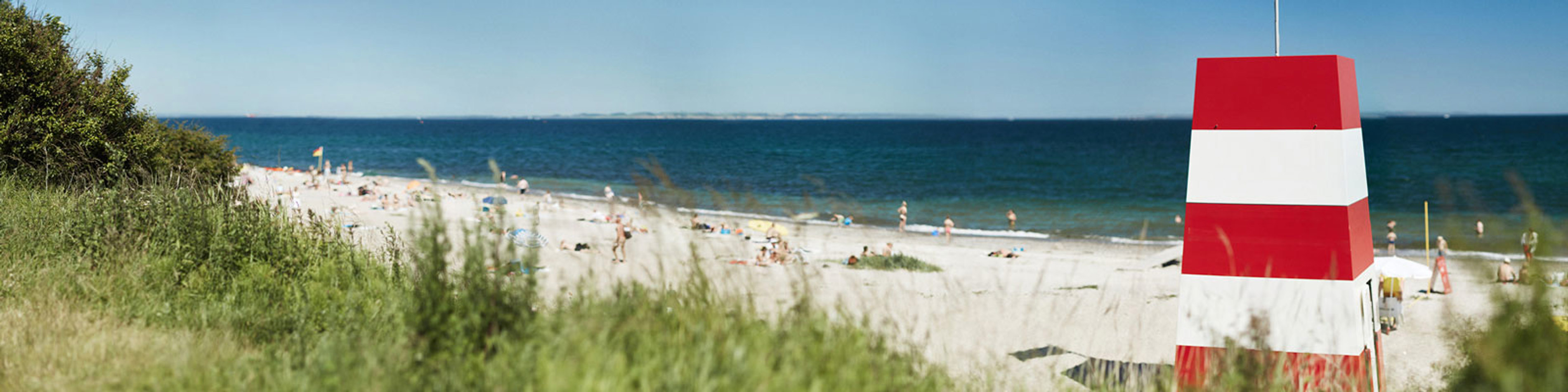 Rød-hvidt fyrtårn ved stranden med udsigt over havet på en solskinsdag, grønne buske i forgrunden.