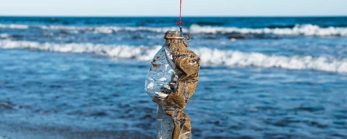 En plastflaska täckt av sand hänger från en lina vid havet med vågor synliga i bakgrunden.