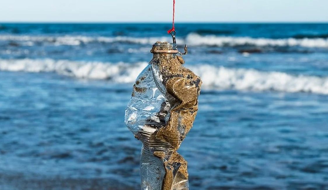 En plastflaska täckt av sand hänger från en lina vid havet med vågor synliga i bakgrunden.