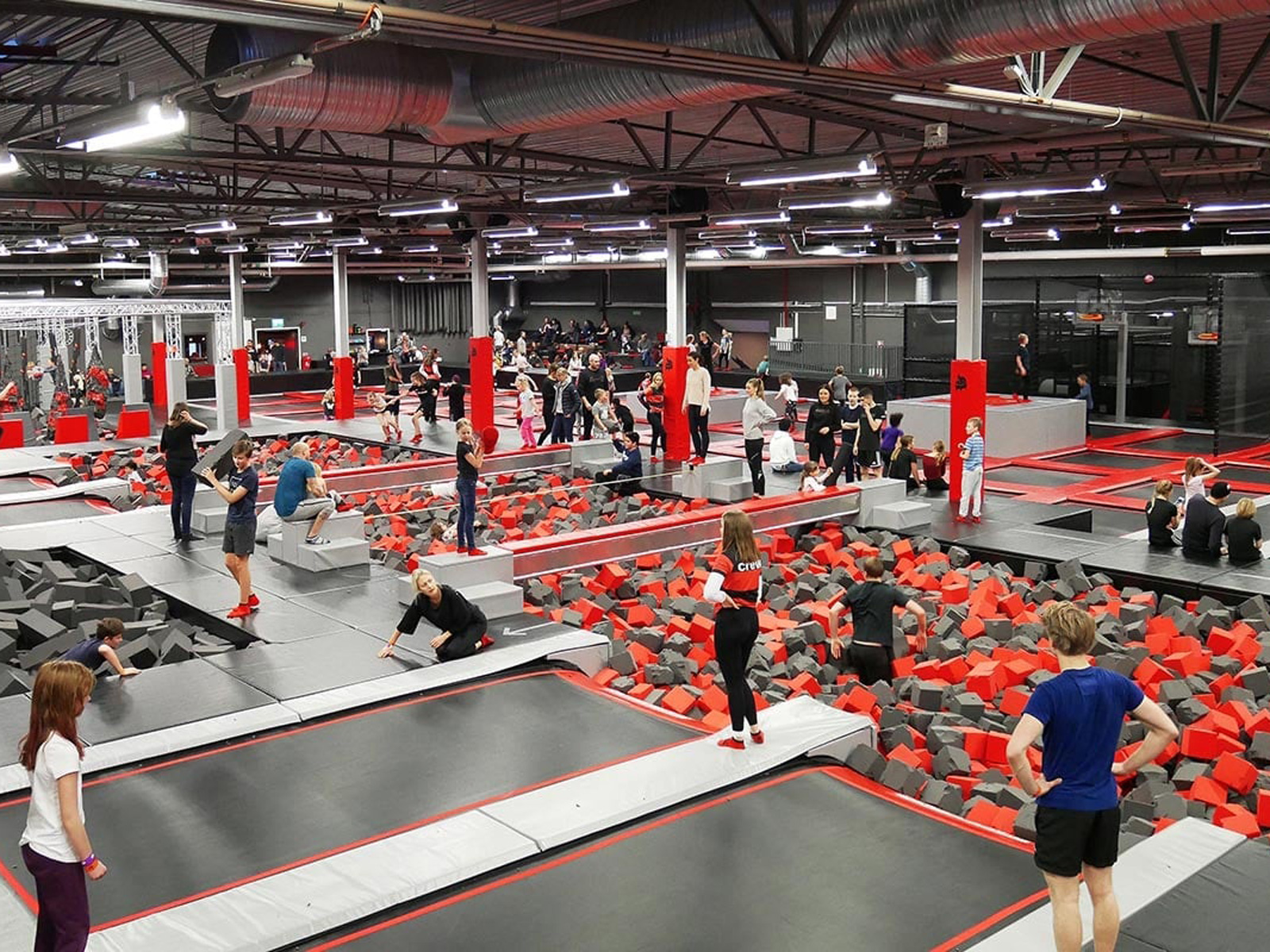 Indendørs trampolinpark med mange børn og voksne som hopper og leger på skumfyldte trampolinområder.