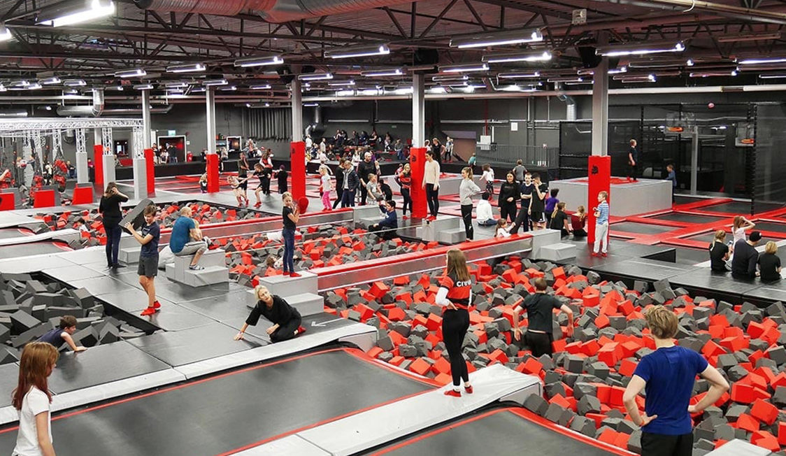 Indendørs trampolinpark med mange børn og voksne som hopper og leger på skumfyldte trampolinområder.